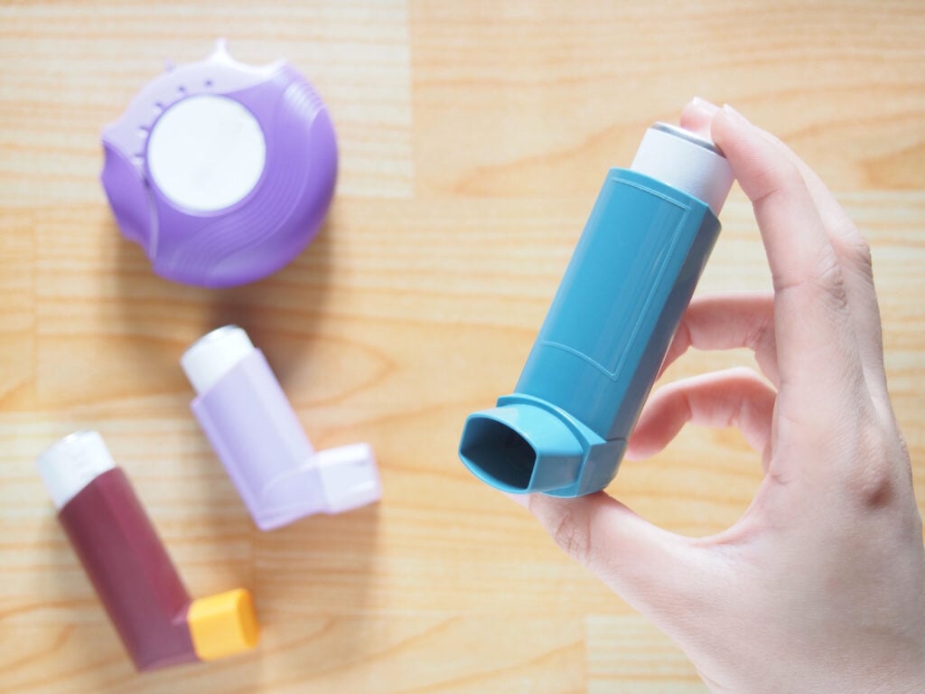 Patient's hands holding blue asthma inhaler with set of asthma/COPD inhalers on wood table. Pharmaceutical products for treat lung inflammation and relief asthma attack. Health and medical concept.